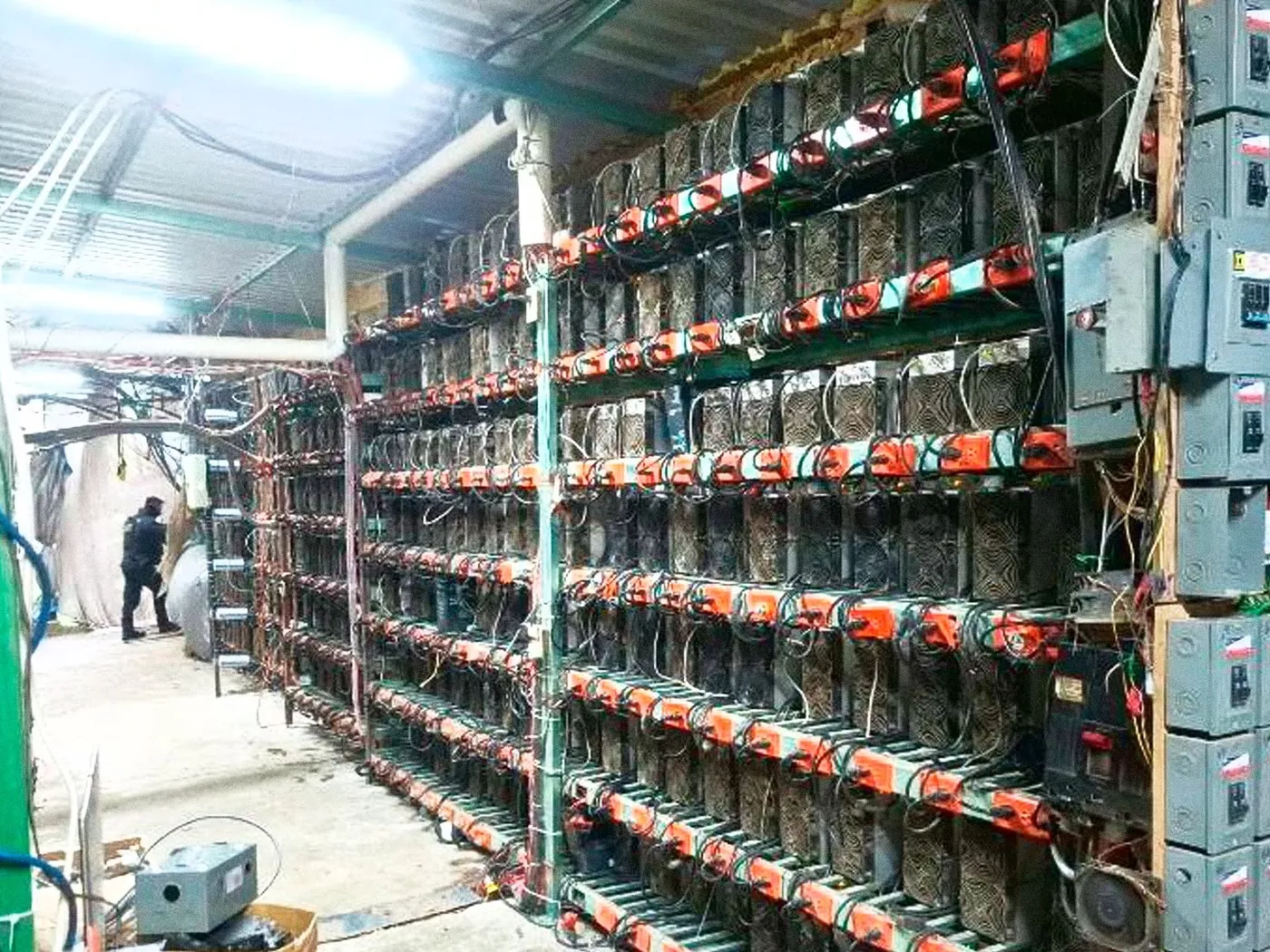 Rows of crypto mining machines on metal racks with orange power connectors and grey breaker boxes inside a corrugated-roof building in Tlaola, Puebla