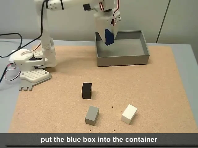 A small white 3D-printed robot arm, the SO-101, mounted at the edge of a cork board on a grey table. Its gripper holds a blue box above a grey open tray at the back of the board. A black cube, a grey cube and a cream cube sit on the cork in front of it. A caption bar along the bottom reads put the blue box into the container.