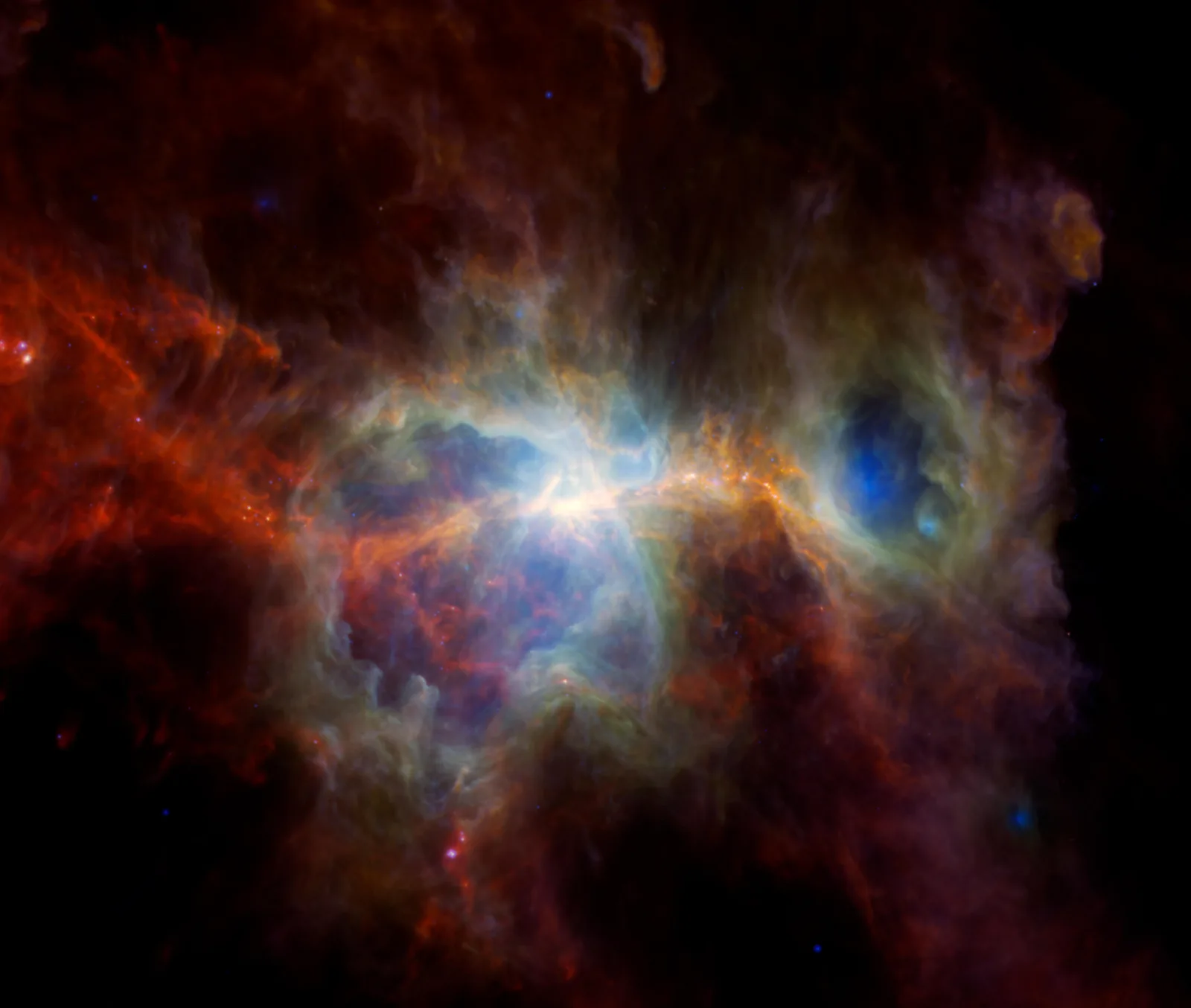 The Orion Nebula in infrared light: two dark blue cavities carved by massive stars sit in a bright white and blue core, ringed by green and orange dust, with red filaments of cold dust spreading across the left and bottom of the frame against black space.