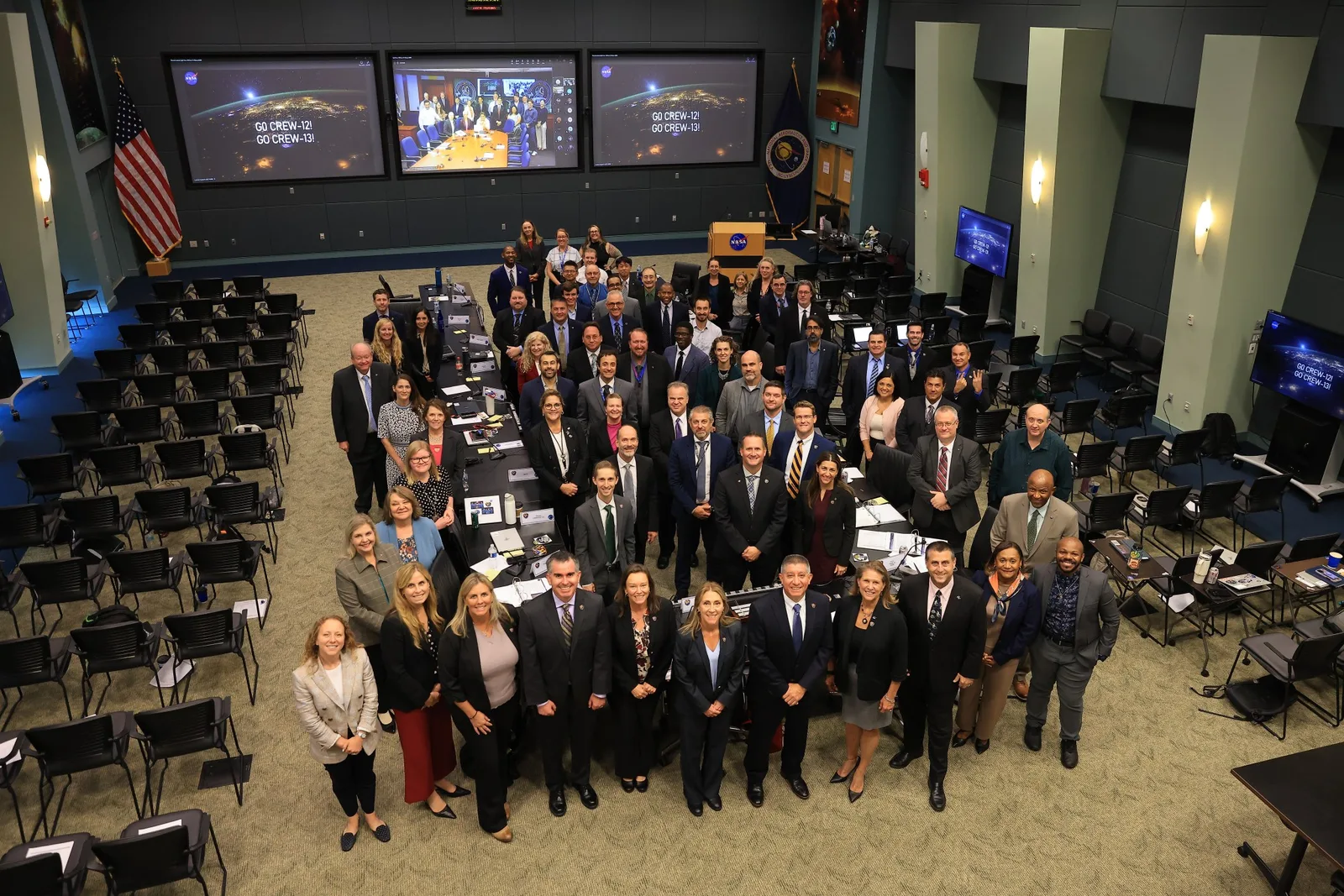 Overhead photograph of dozens of managers from NASA, SpaceX and international partners standing around a long table in a Kennedy Space Center review room, with an American flag, a NASA flag and wall screens reading Go Crew-12! Go Crew-13!