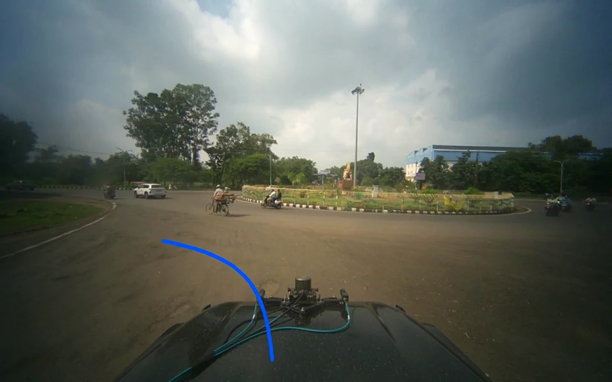 Forward-facing camera view from Odyssey's test car approaching a large roundabout on a public road in India, with motorcycles, a cyclist and a white car crossing ahead, and the driving policy's predicted path drawn as a blue curve across the tarmac.
