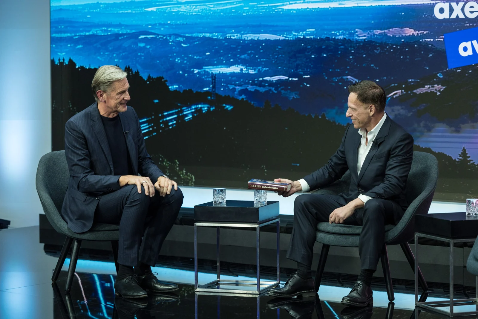 Mathias Döpfner and Peter Thiel sit facing each other in grey armchairs on a stage lit in blue, with a projected night landscape behind them. Thiel, in a dark suit and open-collared shirt, holds out a book with Tolkien on its spine; glasses of water stand on a small table between them.