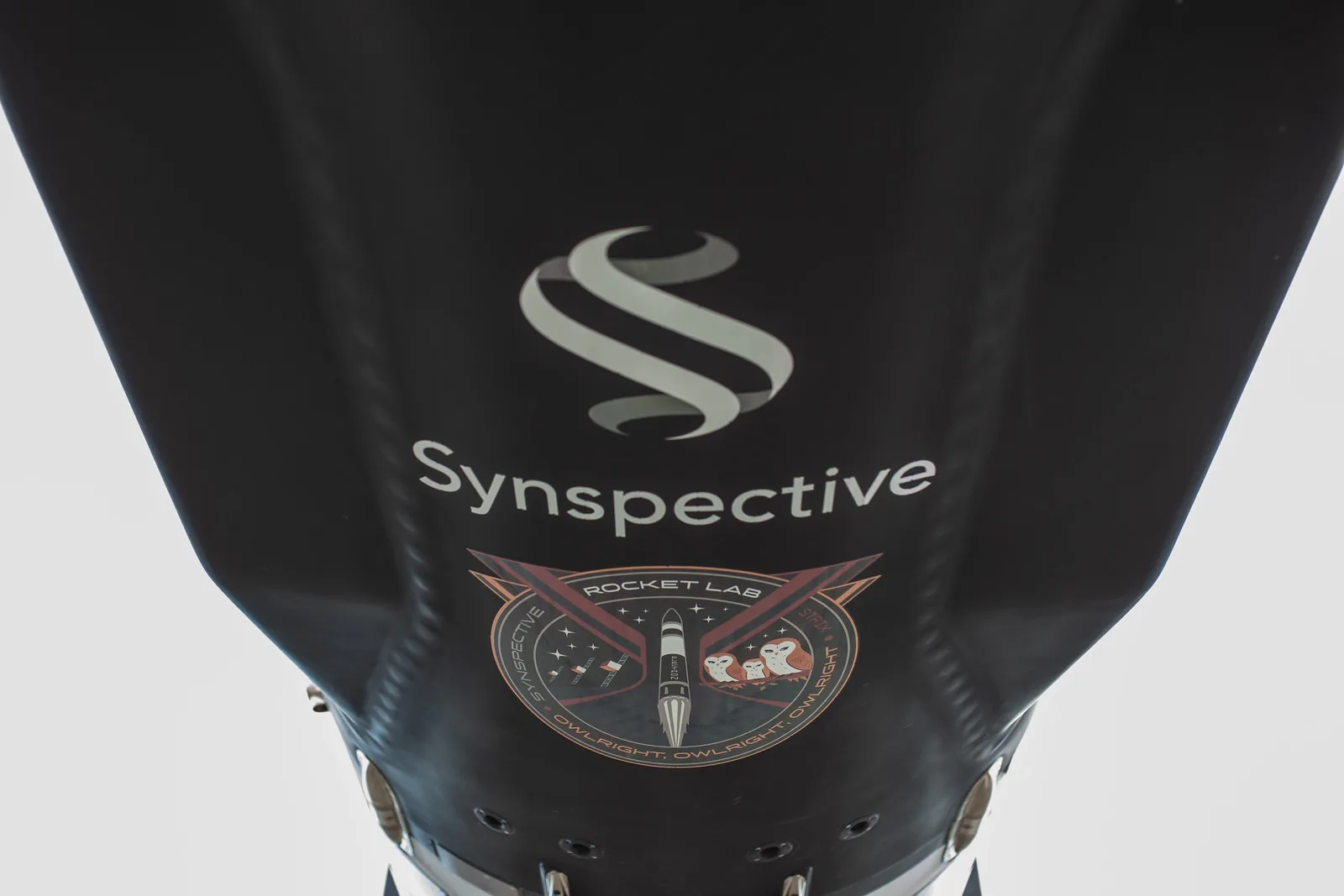 Close view of the black Electron payload fairing, carrying the white Synspective logo, a curved S above the company name, and below it the round mission patch showing an Electron rocket beside three owls, ringed by the words Owlright, Owlright, Owlright.