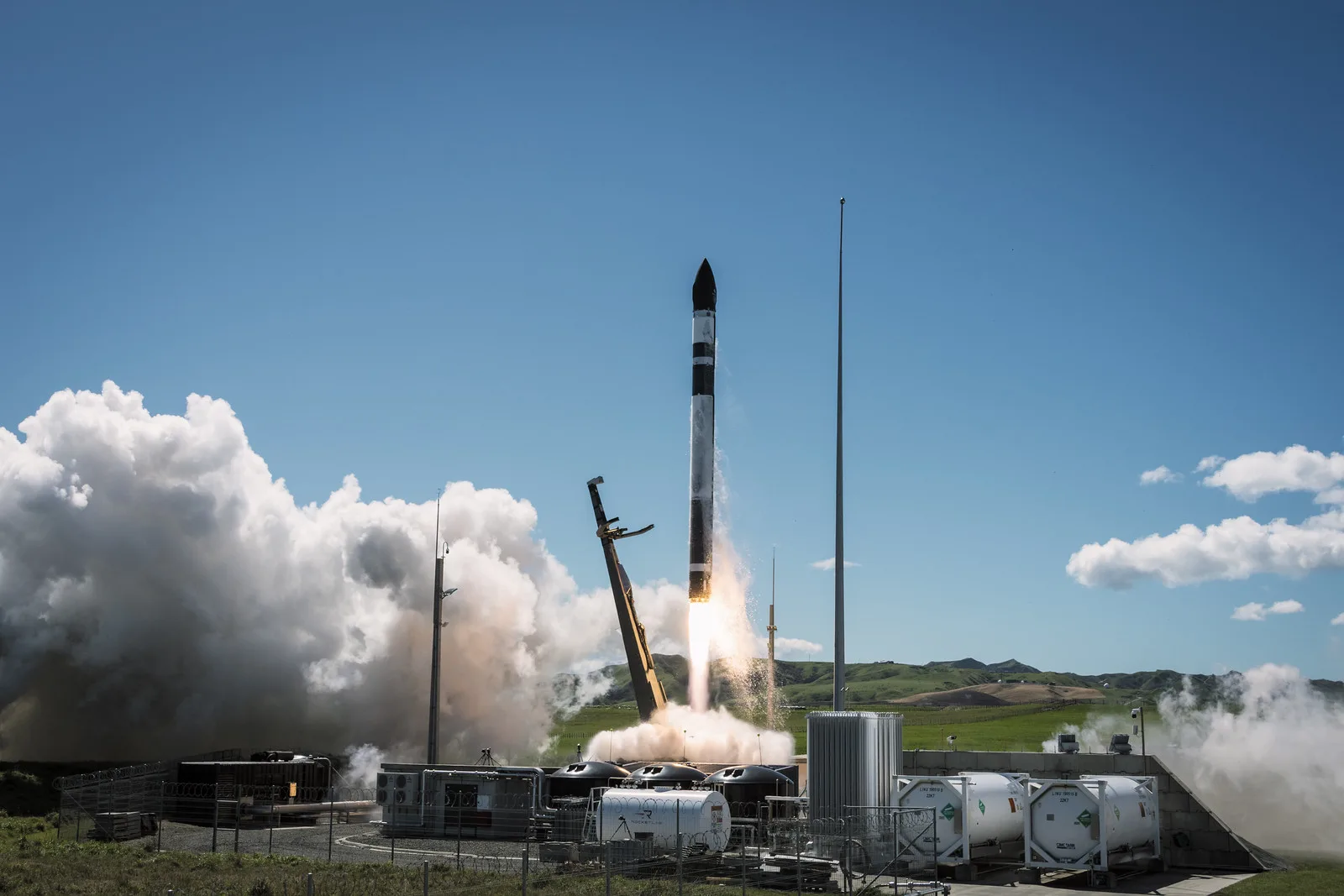 A black and white Electron rocket rising on a bright exhaust flame from Launch Complex 1 on the Mahia Peninsula, with white clouds of steam rolling across the pad, the launch mount's arm swung back, storage tanks in the foreground and green hills under a clear blue sky.