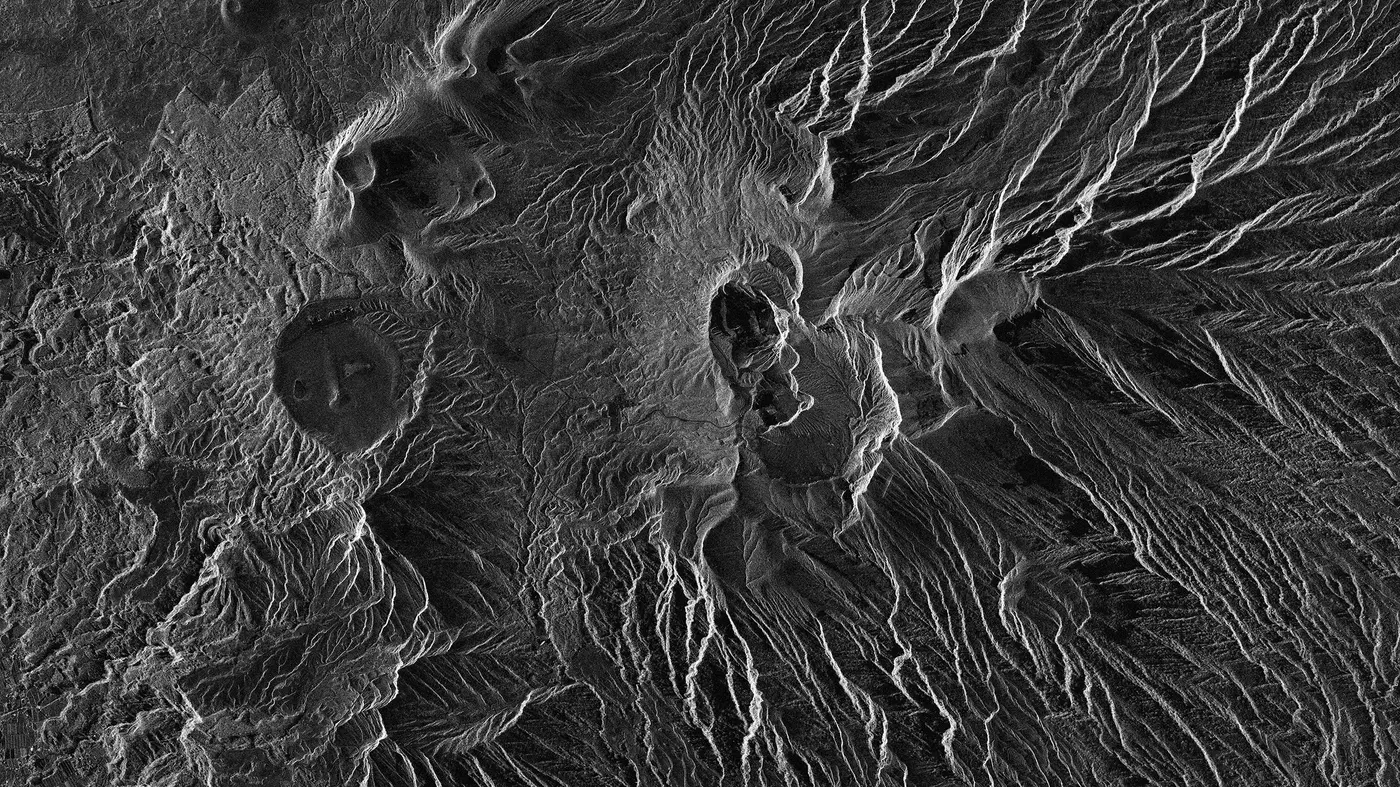 A black and white radar image of Mount Aso in Japan, showing the volcano's craters as smooth dark bowls and hundreds of ridges and gullies radiating down its slopes, lit in fine relief by the radar echoes.