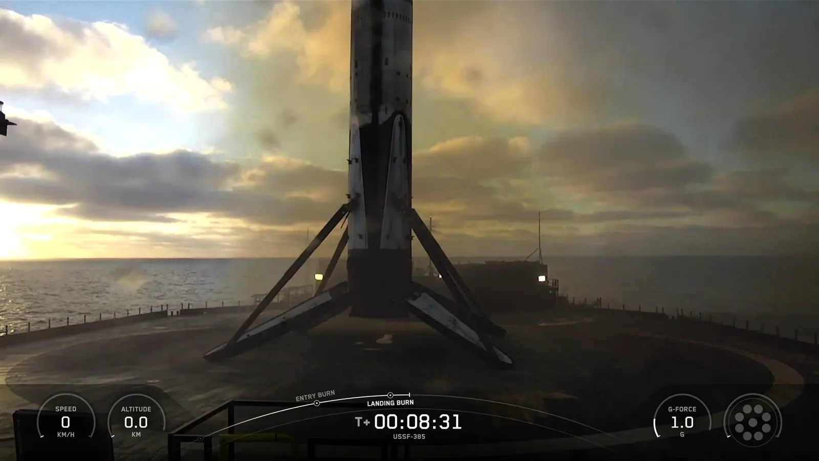 SpaceX webcast frame from the droneship deck: the soot-covered Falcon 9 first stage standing on four landing legs against a cloudy dawn sky over the Pacific, with the webcast clock at T+00:08:31, speed 0 km/h and altitude 0.0 km.