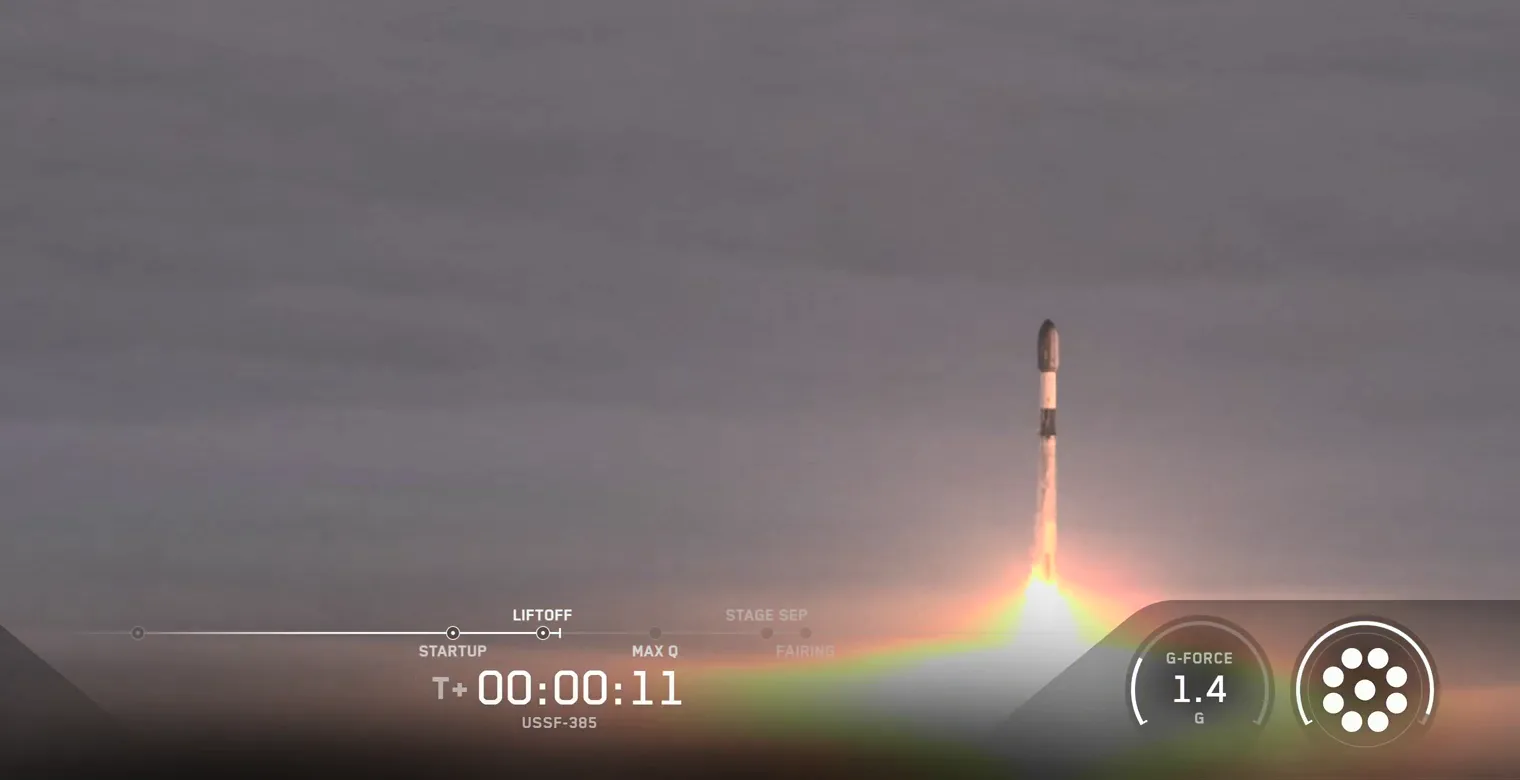 SpaceX webcast frame of Falcon 9 rising above a grey layer of fog on a column of flame, with the webcast clock reading T+00:00:11 and the mission label USSF-385, and a g-force gauge reading 1.4.