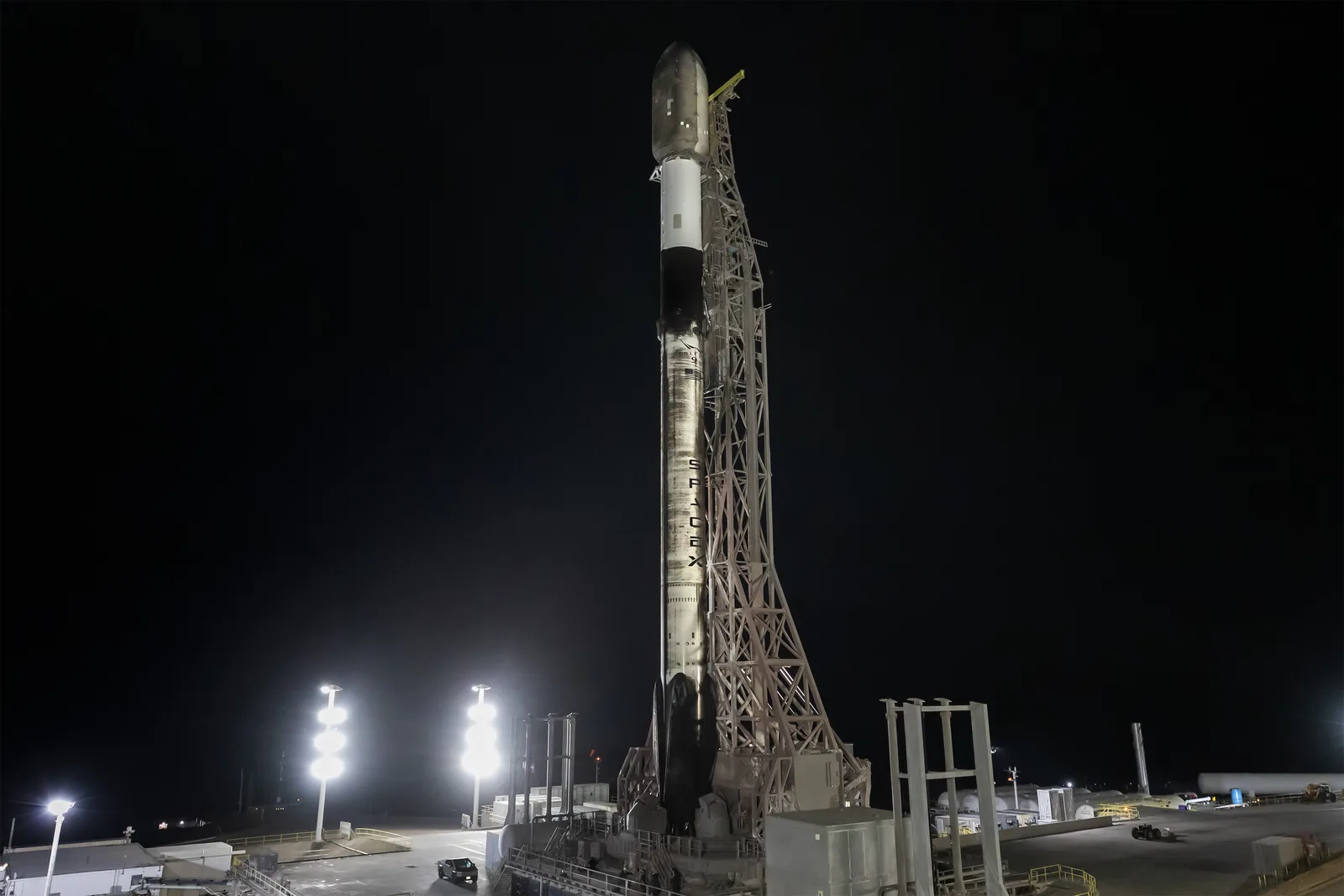 A SpaceX Falcon 9 on its launch mount beside the steel tower at Space Launch Complex 4E, Vandenberg, lit by floodlights against a black pre-dawn sky, its first stage streaked with soot and the payload fairing on top.