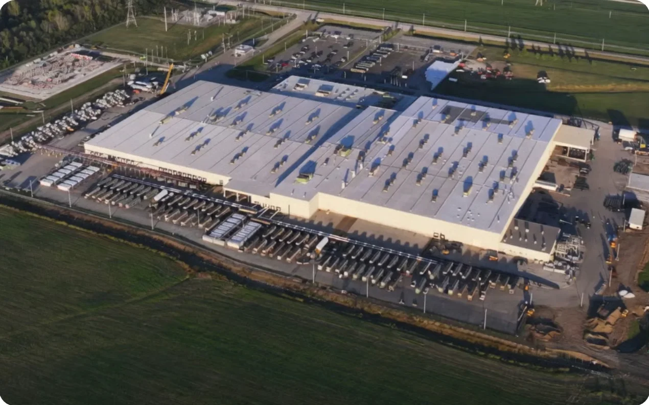 Aerial photograph of the Colossus 1 data centre in Memphis: a long pale single-storey building with rows of rooftop units, surrounded by car parks, lines of equipment trailers and green fields, with an electrical substation at the top left.