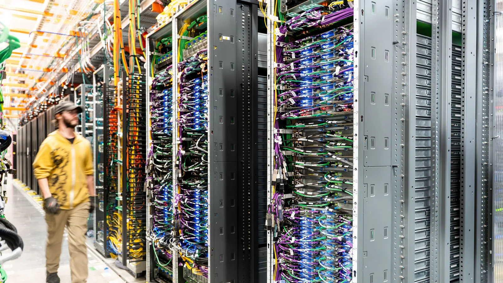 Photograph of an AWS engineer in a yellow jacket and cap walking along an aisle of tall black server racks, their fronts packed with blue, purple and green network cables, under yellow cable trays and bright overhead lights.