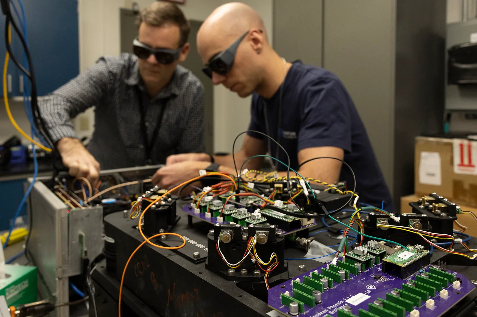 Two Quantum Brilliance engineers in dark laser-safety glasses lean over an open optical bench at Oak Ridge National Laboratory, adjusting orange, yellow and blue cables that run between black optical mounts and purple circuit boards studded with green connectors.