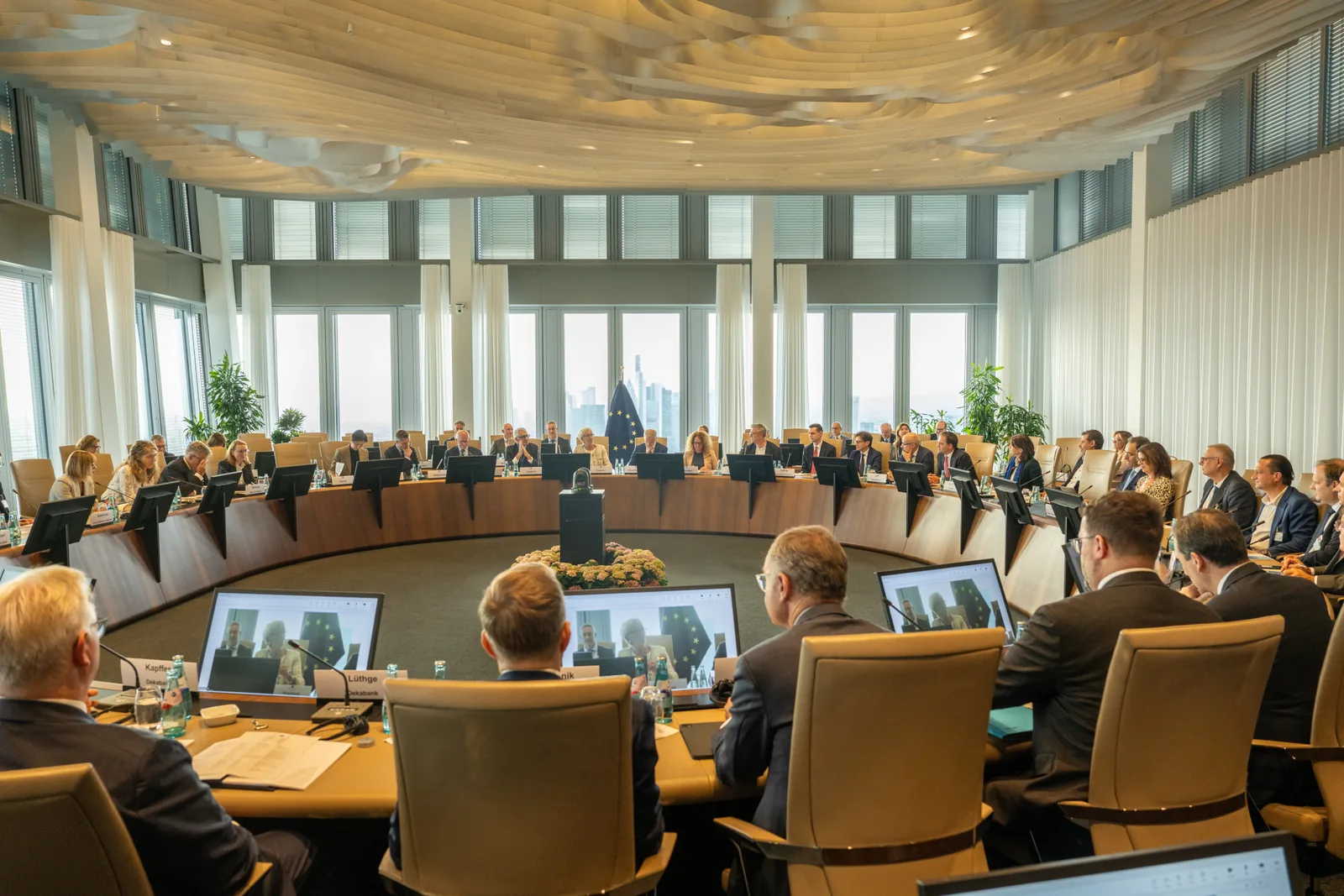 A wide photograph of a round meeting room at the ECB in Frankfurt during the Pontes go-live event. Several dozen people sit at a large curved wooden table with screens and microphones in front of them, a European Union flag stands at the far side, and floor-to-ceiling windows behind it look out over the Frankfurt skyline. In the foreground, participants seen from behind face laptops showing a video call.