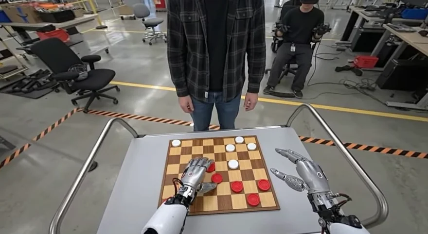 A Genie 4 simulation seen from the robot's point of view: two silver robotic hands rest on a wooden checkerboard of red and white pieces on a metal cart, with a person in a checked shirt standing opposite in a robotics workshop with chairs and yellow-and-black floor markings.