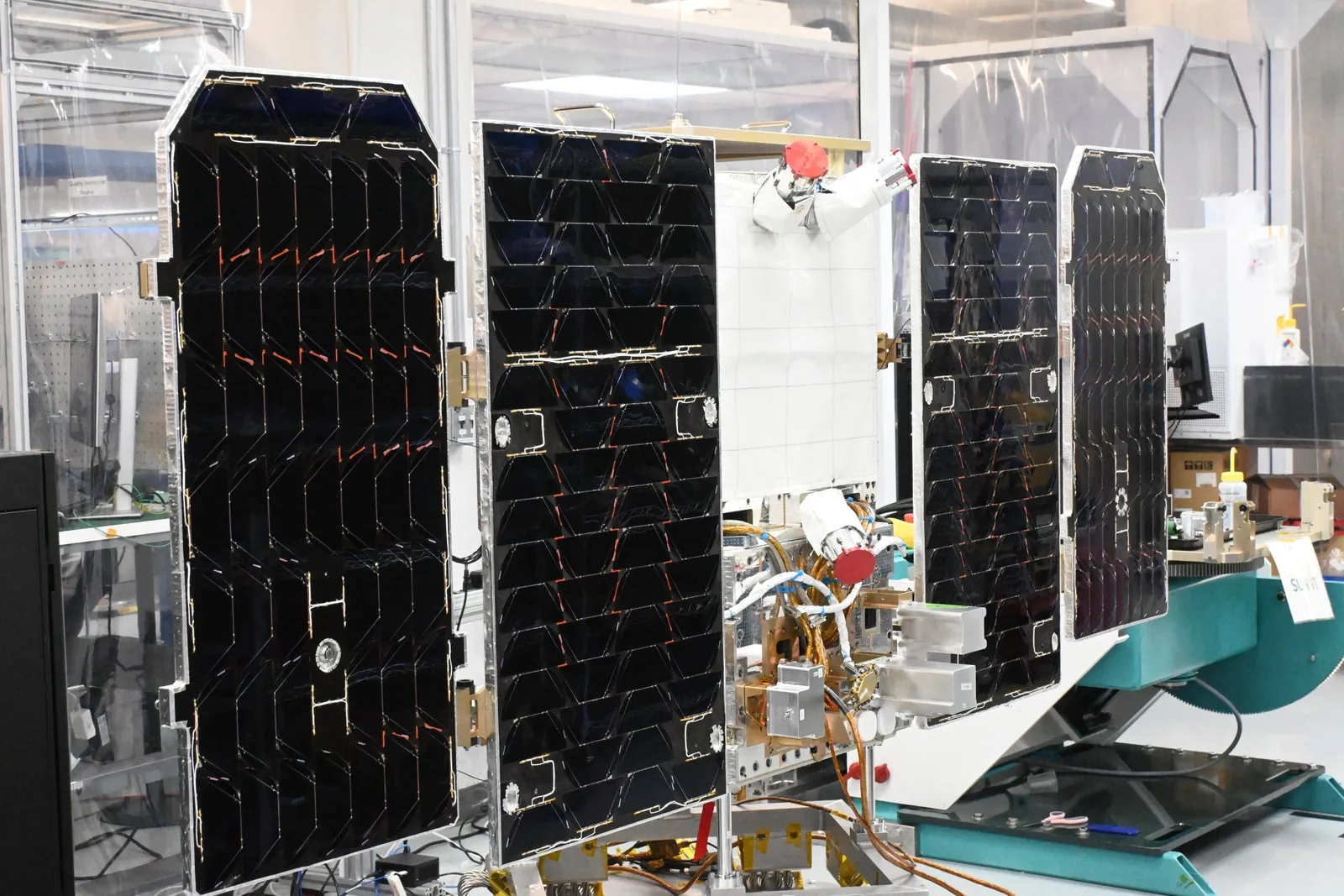 The Project Suncatcher prototype satellite in a clean room: four tall black solar panels folded out on hinges, two either side of a white central body, with gold cabling, metal instrument boxes and two red protective caps, mounted on a teal handling stand.