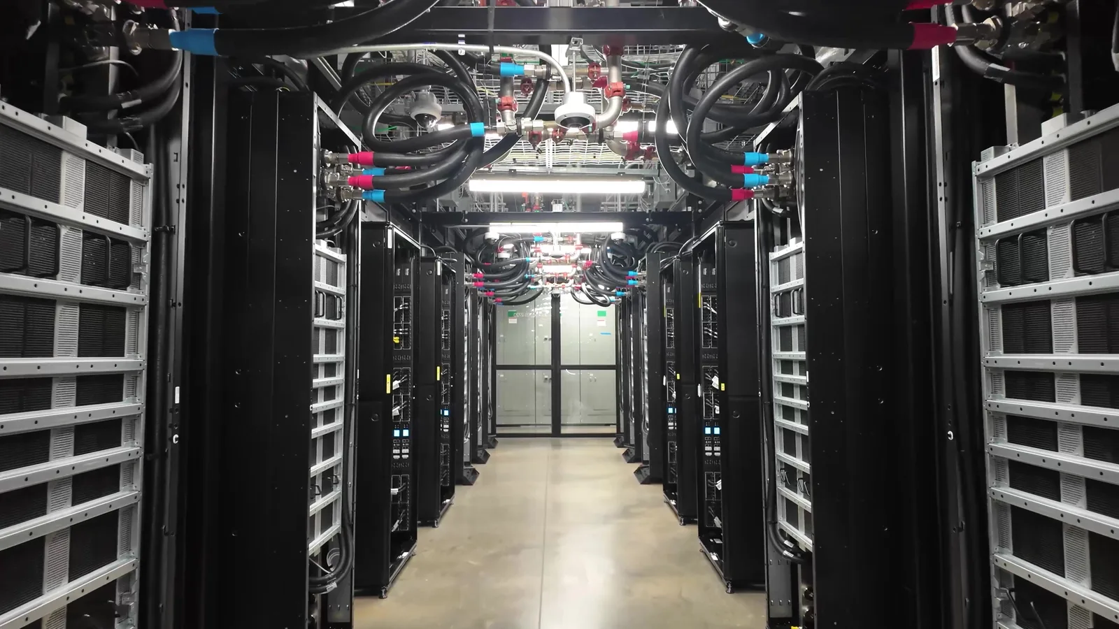 Photograph looking down a long aisle between rows of black server racks in an Oracle data hall, with thick black hoses fitted with red and blue connectors running overhead and grey electrical cabinets at the far end.