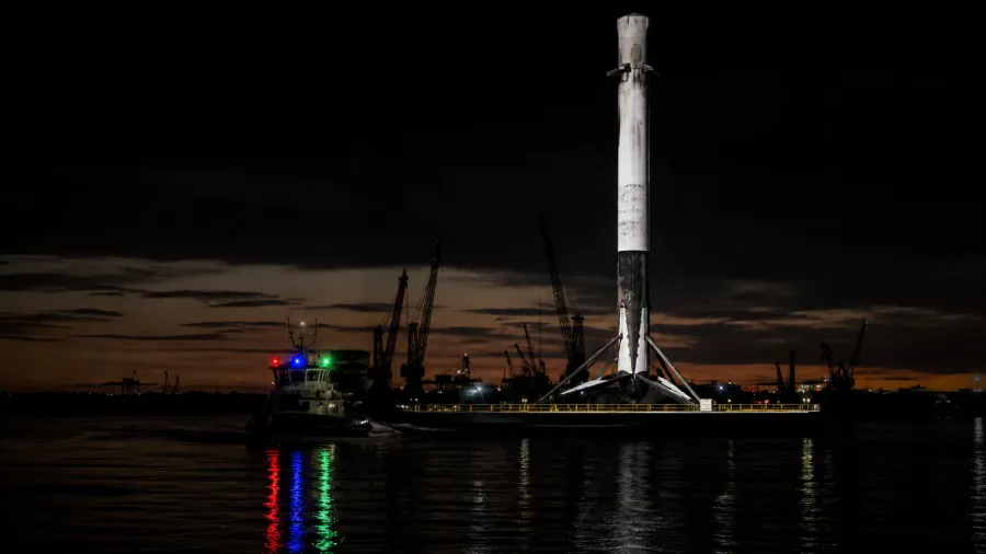 Concept artwork of a recovered booster standing on a barge towed towards a dark port at dusk, tug lights in red, blue and green.